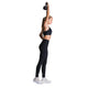 En kvinde i Fusion X-Long Training Tight, Dame, SuperBlack og en sort sports-bh står oprejst på en almindelig hvid baggrund og holder en kettlebell over hovedet. Hun fuldender looket med svedtransporterende leggings og hvide sneakers.