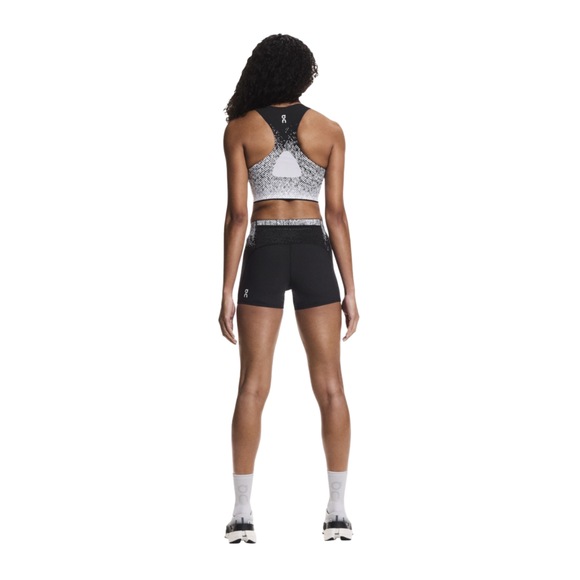 En person med krøllet hår, set bagfra, har en sort og hvid On Zero Short Tights Dame på, en matchende sports-bh, hvide strømper og hvide sneakers. De svedtransporterende tights holder det sporty look frisk mod den ensfarvede baggrund.