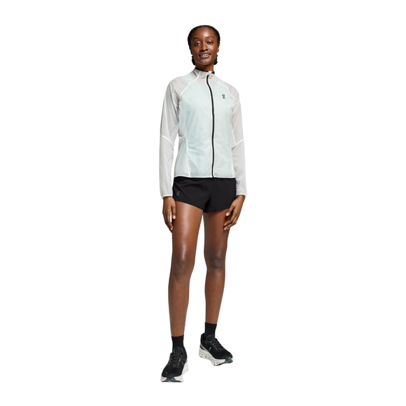 En person står med front mod en hvid baggrund iført On Zero Jacket til kvinder i hvid, sammen med sorte shorts, sorte sokker og sorte sportssko.