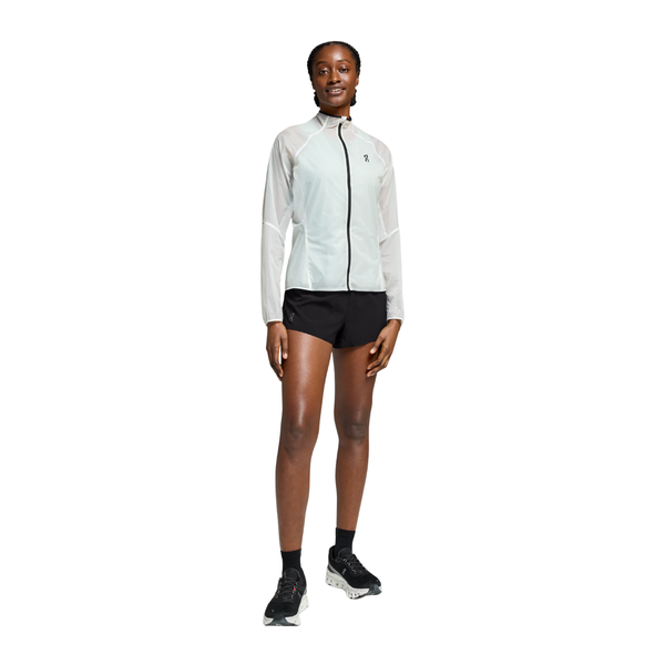 En person står med front mod en hvid baggrund iført On Zero Jacket til kvinder i hvid, sammen med sorte shorts, sorte sokker og sorte sportssko.