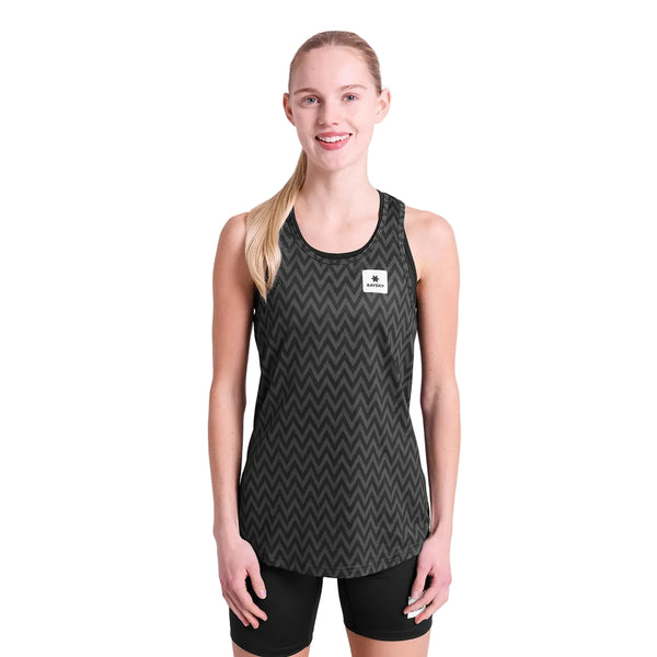 En person med langt blondt hår bundet tilbage smiler fremad, iført Saysky Zig Zag Combat Singlet, Dame, Black Zigzag, med sorte shorts på en almindelig hvid baggrund.