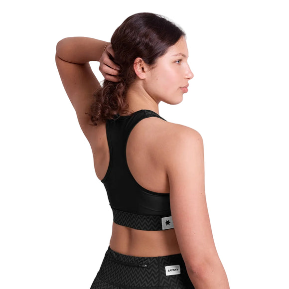 En kvinde med brunt hår i hestehale står med ryggen til og venstre hånd på hovedet. Hun er iført Saysky Zig Zag Combat Sports Bra, Dame, Black Zigzag og sorte mønstrede shorts - ideel til højintensivt løb med genanvendte Combat-materialer.