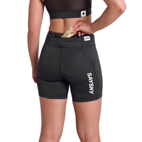 En kvinde iført Saysky Zig Zag Combat+ Short Tights 7" i Black Zigzag, fremstillet af genbrugspolyester, ses bagfra i færd med at lægge en energigel i lommen på linningen. Både hendes top og shorts har Saysky-branding.