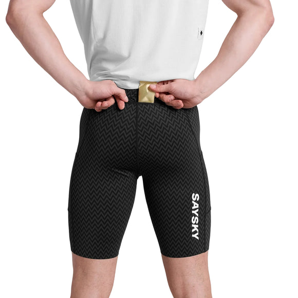 En person i hvid skjorte og sorte Saysky Zig Zag Combat+ Short Tights 9", Herre, Black Zigzag, fremstillet af genbrugspolyester, ses bagfra lægge en guldgenstand i den bageste linningslomme.