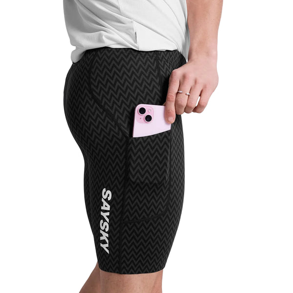 En mand i sorte Zig Zag Combat+ Short Tights 9" fra Saysky med zigzagmønster og "SAYSKY" på siden lægger en pink telefon i lommen, mens han har en hvid skjorte på. Strømpebukserne er lavet af genbrugspolyester.
