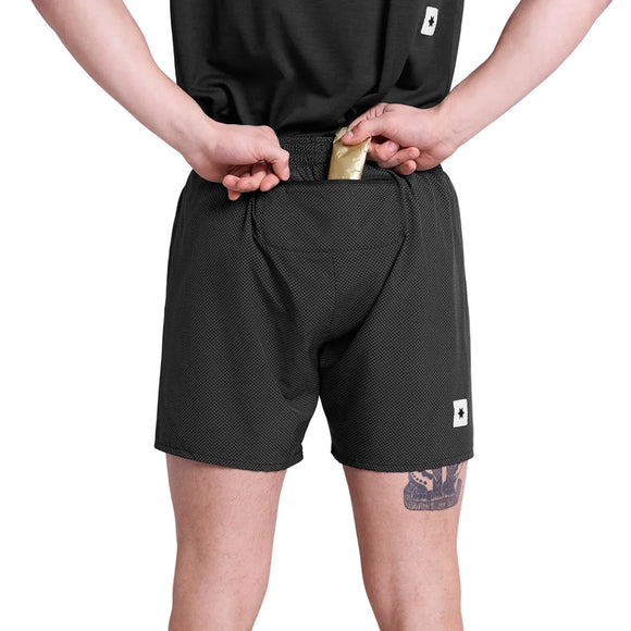 En person iført Saysky Zig Zag Pace Shorts 5" i Black Zigzag ses bagfra i færd med at lægge en guldpakke i shortsens lynlåslomme i linningen. En tatovering er synlig på vedkommendes venstre lår.