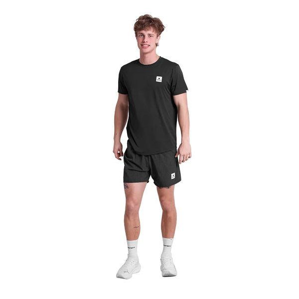 En person står med ansigtet fremad i en sort t-shirt og Saysky Zig Zag Pace Shorts 5", Herre, Black Zigzag, med hvide sokker og sneakers på en hvid baggrund.