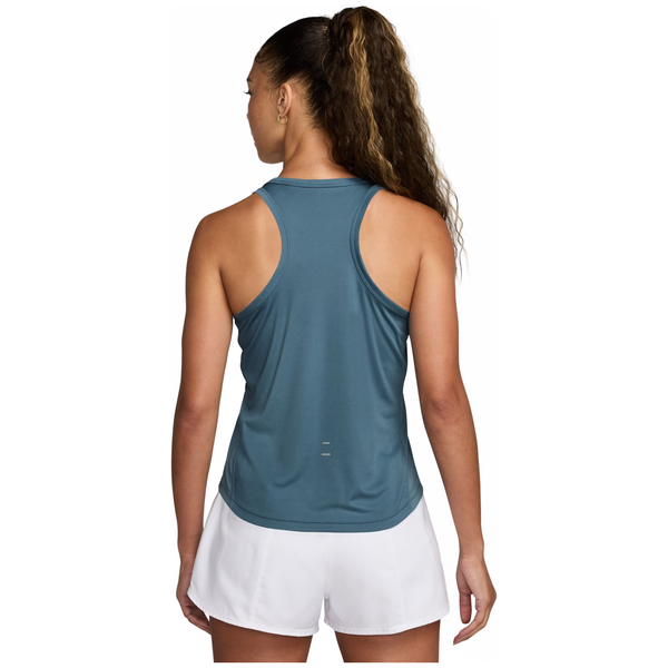 En kvinde med langt krøllet hår i en hestehale kigger væk, iført en Nike Dri-Fit Running Tank Top for dame i Smokey Blue/Reflective Silver og hvide shorts på en almindelig hvid baggrund.