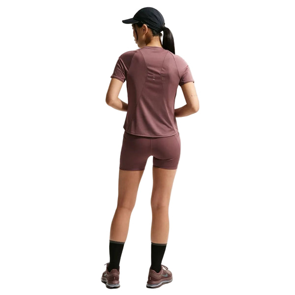 En person vender ansigtet væk, iført mørk kasket, kortærmet lilla sportstrøje, Nike Nk Df Swift Hr 4In Short Dame i Tattoo/Reflective Silv, sorte sokker og lilla sneakers. Baggrunden er helt hvid.