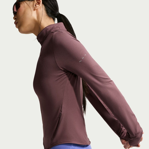 Iført Nikes Swift UV Half Zip Running Top til kvinder i Tattoo/Reflective Silver strækker en person med solbriller sig med hovedet drejet til siden og håret bundet tilbage. Den ensfarvede baggrund fremhæver deres atletiske stil.