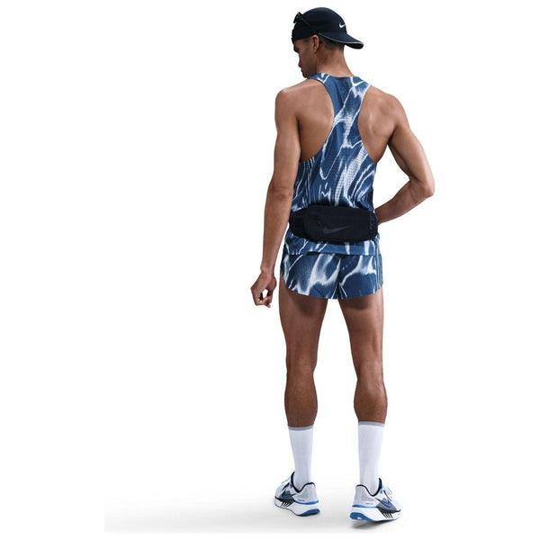 En mand kigger væk iført en Nike Aeroswift Singlet, Herre, Coastal Blue/White løbevest og shorts, hvide sokker, løbesko, en sort bæltetaske og en sort bagudvendt kasket på en hvid baggrund.