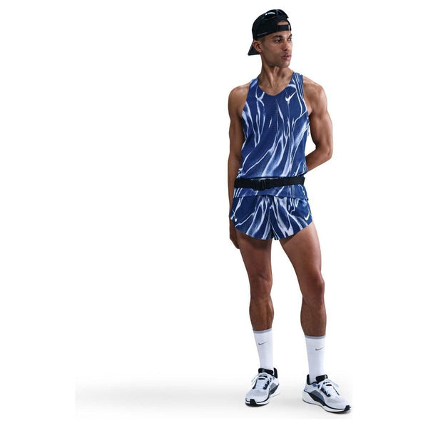 En mand står på en hvid baggrund iført Nike Aeroswift Singlet, Herre, i Coastal Blue/White. Han kombinerer den med matchende shorts, hvide sokker og løbesko og en bagudvendt sort kasket med den ene hånd bag ryggen.