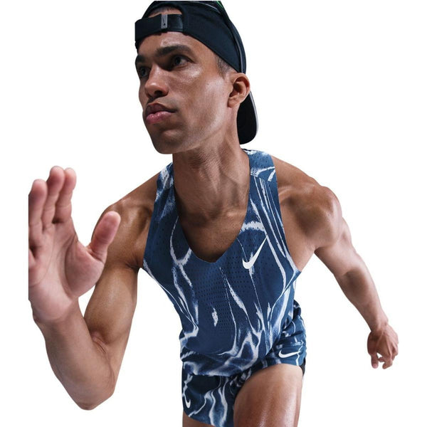 En mand i en Nike Aeroswift Singlet til mænd, Coastal Blue/White, og en bagudvendt sort kasket løber dynamisk mod en hvid baggrund.