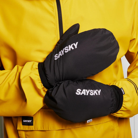 En person iført en gul jakke og Saysky Blaze-luffer - sorte unisex-luffer med "SAYSKY" trykt i hvidt - står med hænderne krydset ved brystet.