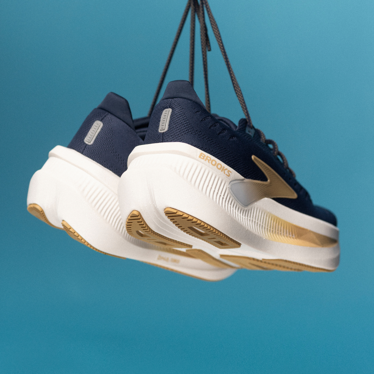 Et par marineblå og hvide løbesko med gulddetaljer hænger i snørebåndene mod en ensfarvet lyseblå baggrund.