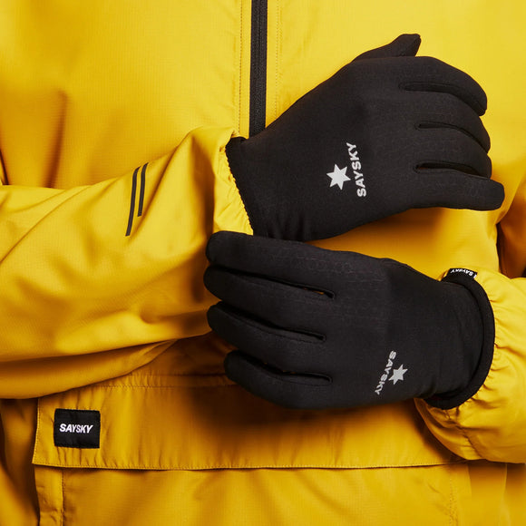 En person i gul jakke bærer sorte Saysky Combat Gloves med skridsikkert greb. Hænderne er krydset i taljen, så man kan se Saysky-logoet på både handskerne og jakken.