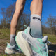 Nærbillede af en person udendørs på græs iført lysegrå løbesko med mintfarvede detaljer og Feetures Elite Light Cushion Mini Crew unisex-sokker med CPH-tekst. I baggrunden ses nøgne træer og blå himmel.