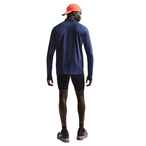 En mand ser væk i en blå langærmet skjorte, sorte shorts over Nike Nk Df Stride Half Tight, Herre, Black/Reflective Silv, brune løbesko, rød kasket og mørke sokker mod en almindelig hvid baggrund.