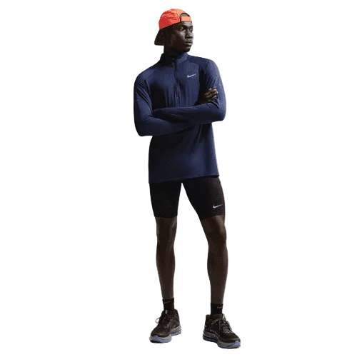 En person står med armene over kors iført en marineblå langærmet top, Nike Nk Df Stride Half Tight shorts i sort/reflekterende sølv, sorte løbesko og en rød baglæns kasket. Nike-logoer er synlige på tøjet mod en almindelig hvid baggrund.