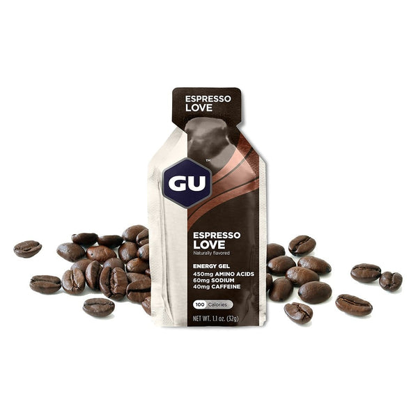 En pakke GU Energy Gel, Espresso Love fra GU Energy står oprejst blandt kaffebønner på en hvid baggrund og viser sine ingredienser og næringsoplysninger på pakken som et sportsernæringsprodukt.