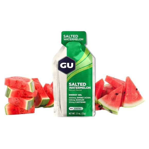 En pakke GU Energy Gel, Salted Watermelon fra GU Energy står oprejst blandt friske vandmelonskiver på en hvid baggrund. Den grønne og sølvfarvede emballage, der er mærket som en kulhydratgel, fremhæver dens ernæringsmæssige detaljer.