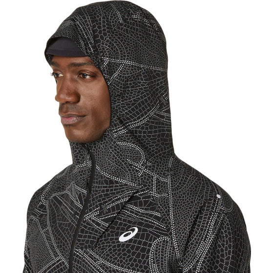 En person i ASICS Limited Series Lite-Show Jacket, Herre, Performance Black - en sort løbejakke med et hvidt geometrisk mønster, Asics-logo på brystet og ASICS-refleks-teknologi- ser lidt til venstre mod en almindelig baggrund.