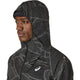 En person i ASICS Limited Series Lite-Show Jacket, Herre, Performance Black - en sort løbejakke med et hvidt geometrisk mønster, Asics-logo på brystet og ASICS-refleks-teknologi- ser lidt til venstre mod en almindelig baggrund.