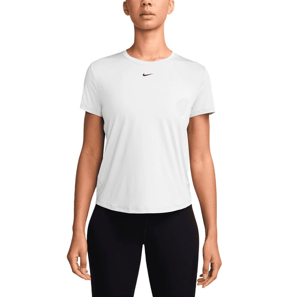 En kvinde står med front mod en hvid baggrund, klædt i sorte bukser og en Nike Dri-Fit Short-Sleeve Top (Dame) i hvid/sort, fremstillet af genanvendt polyester med et lille sort Nike-logo på brystet.