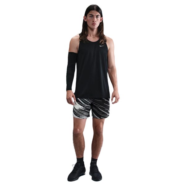 En person med langt hår ser fremad, iført en sort ærmeløs top, Nike Nk Df Stride 7In Bf Shrt Op2 løbeshorts i Photon Dust/Reflective Silver, et sort ærme på højre arm, sorte sokker og sorte sportssko. Baggrunden er helt hvid.