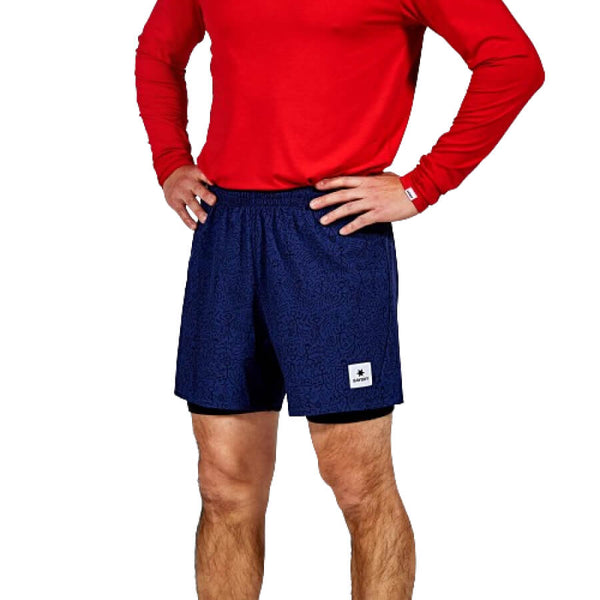 En person står med hænderne på hofterne iført en langærmet rød skjorte og Saysky CC Pace 2 in 1 Shorts 5", Herre, Blue. Shortsene har en lille hvid plet på venstre ben. Hovedet er ikke synligt på billedet.
