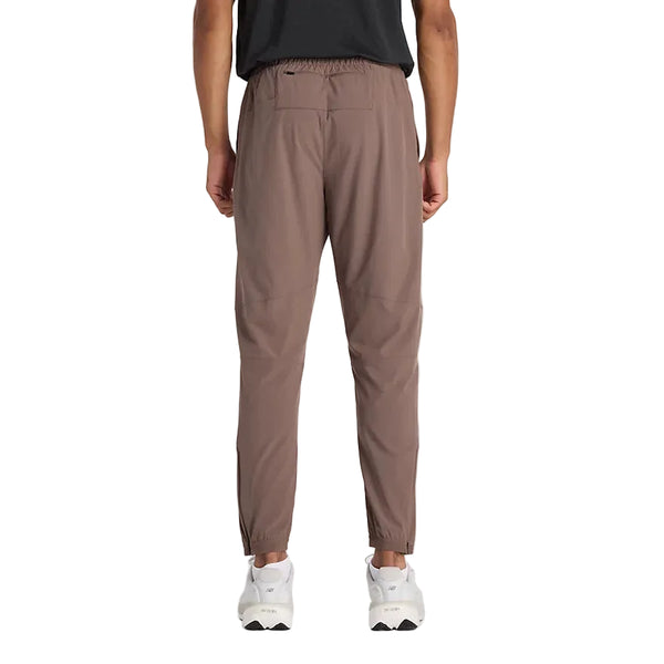 En person ses bagfra iført sort skjorte, New Balance Athletics Run Jogger Herre Cortado med elastisk linning og manchetter og hvide sportssko, stående på en almindelig hvid baggrund.