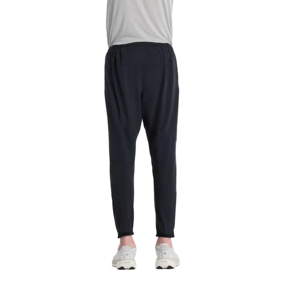 En person iført en lysegrå skjorte, New Balance Ath Run Jg løbetight til mænd i sort og hvide sneakers står med ryggen til kameraet mod en almindelig hvid baggrund.