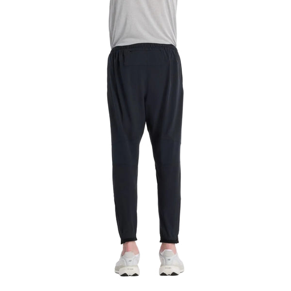 En person iført en lysegrå skjorte, New Balance Ath Run Jg løbetight til mænd i sort og hvide sneakers står med ryggen til kameraet mod en almindelig hvid baggrund.