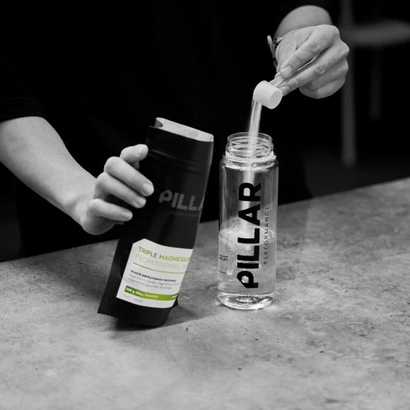 En person hælder en skefuld pulveriseret kosttilskud fra en Pillar Performance Triple Magnesium-pakke i en 500 ml BPA-fri Pillar Performance Micro Shaker på en bordplade.