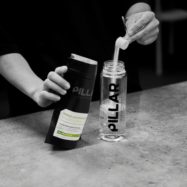 En person hælder en skefuld pulveriseret kosttilskud fra en Pillar Performance Triple Magnesium-pakke i en 500 ml BPA-fri Pillar Performance Micro Shaker på en bordplade.