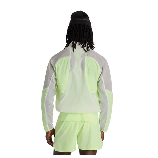 En person med langt hår iført sort kasket, New Balance Rc Jacket, Herre, Afterglow i lysegrøn og grå og matchende shorts står med ryggen til kameraet på en hvid baggrund.