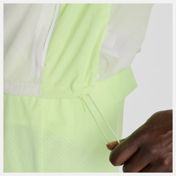 En person justerer snoren på lysegrønne perforerede shorts, mens han er iført en hvid top og New Balance RC Jacket, Herre, Afterglow. Kun underkroppen og den ene hånd er synlige.