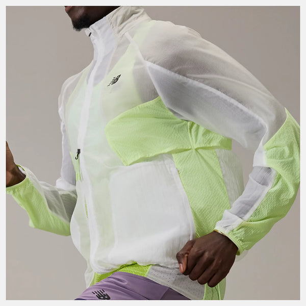 En mand iført en hvid og lysegrøn New Balance Rc Jacket, Herre, Afterglow, parret med lilla shorts, er fanget midt i en løbestilling mod en almindelig baggrund.