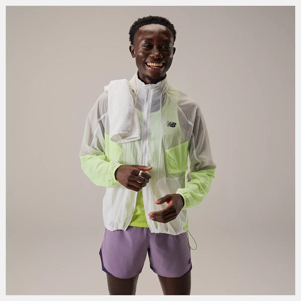 En person står på en almindelig baggrund, smilende og iført en let New Balance Rc Jacket, Herre, Afterglow, lilla løbeshorts og et hvidt håndklæde over skulderen.