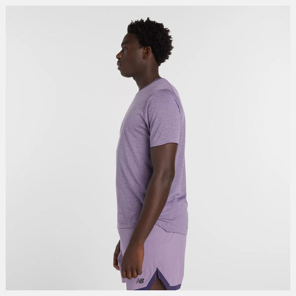 En person står i profil mod venstre, iført en New Balance Athletics T-shirt, Herre, i Candied Violet Heather med synligt logo. Baggrunden er almindelig hvid.