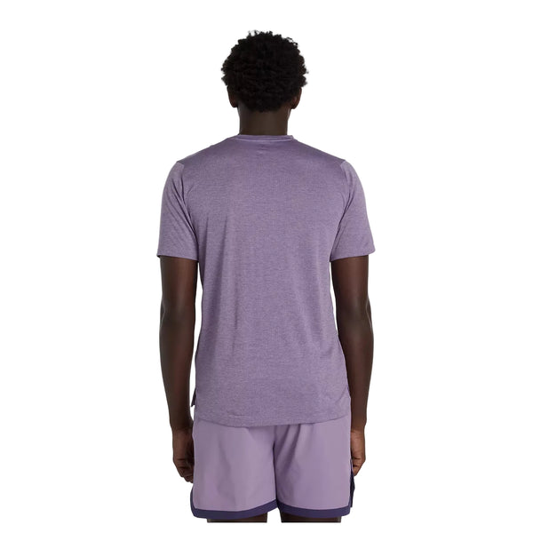 En person står med ryggen til kameraet iført en New Balance Athletics T-shirt til mænd i Candied Violet Heather og lyslilla sportsshorts på en almindelig lys baggrund.