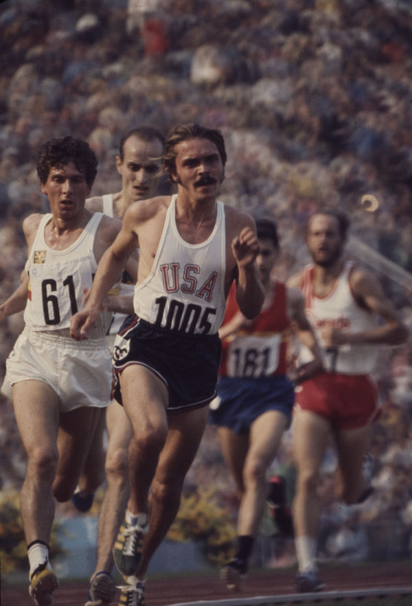 En gruppe mandlige atleter løber på en bane, anført af en løber i USA-uniform med nummeret 1005, under et konkurrenceløb på et overfyldt stadion.