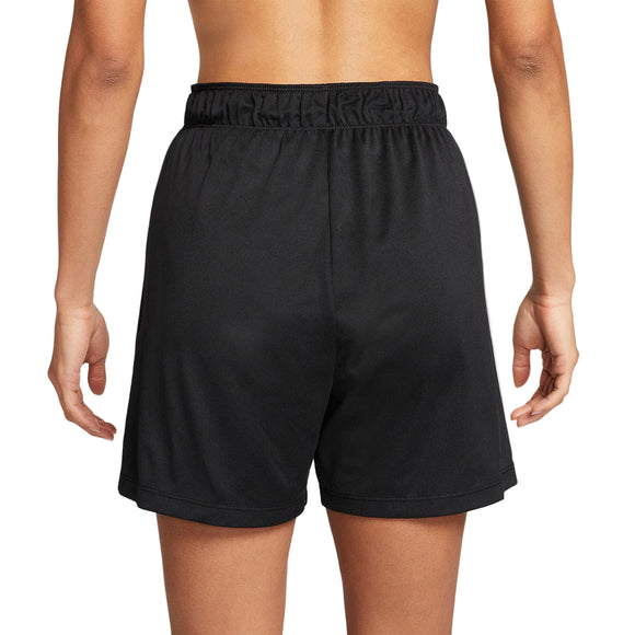 En person står med front mod kameraet i Nike Dri-FIT Fitness Mid-Rise 5" Unlined Shorts til kvinder i Black/Black/White/Reflective Silver med elastisk linning. Armene hviler i siden på en almindelig hvid baggrund.