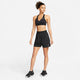 En kvinde står på en hvid baggrund iført Nike Dri-FIT Fitness Mid-Rise 5" Unlined Shorts i Black/Black/White/Reflective Silver og en sort sports-bh, mens hun kigger til siden med den ene hånd på hoften.