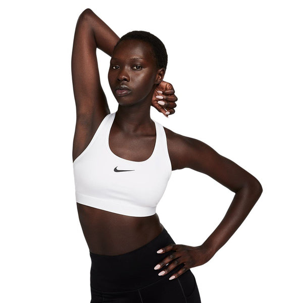 En kvinde poserer med den ene arm bøjet over hovedet iført en hvid Nike Swoosh Medium Sports Bra (Dame, White/Stone Mauve/Black) og sorte leggings på en almindelig hvid baggrund.