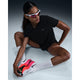 En person i reflekterende solbriller, en sort Nike Dri-FIT Short-Sleeve Running Top (Dame, Black/Reflective Silv), hvide knæhøje fugttransporterende sokker og sportssko med pink og hvide såler sidder på gulvet og rører ved sin sko.