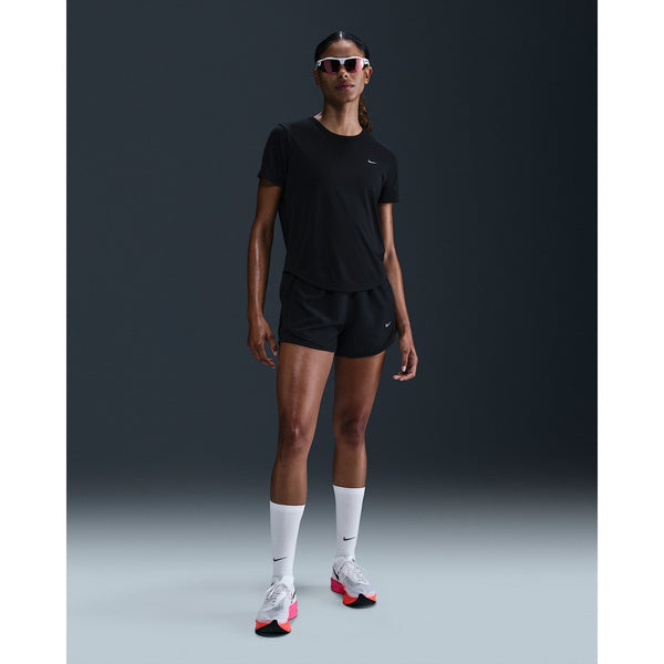 En person står med front mod en almindelig mørk baggrund iført sorte Nike Dri-FIT sportsshorts, en Dri-FIT kortærmet løbetop til kvinder i Black/Reflective Silver fra Nike, hvide knæstrømper, hvide løbesko med pink såler og hvide solbriller.