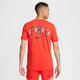 En person kigger væk i en Nike Dri-FIT løbe-T-shirt til mænd i Picante Red med fed sort og hvid "SPORT"-tekst og et Nike-logo. Matchende shorts viser "STOP" på venstre ben og "PRE" på højre.