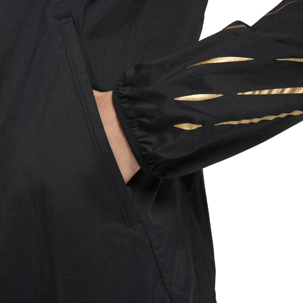 En person iført Nike Nk Rpl Jakob Jkt, Herre, i Black/Black/Metallic Gold har hånden i sidelommen. Det beskårne billede viser kun overkroppen og armen og fremhæver gulddetaljer på ærmet.