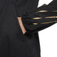 En person iført Nike Nk Rpl Jakob Jkt, Herre, i Black/Black/Metallic Gold har hånden i sidelommen. Det beskårne billede viser kun overkroppen og armen og fremhæver gulddetaljer på ærmet.